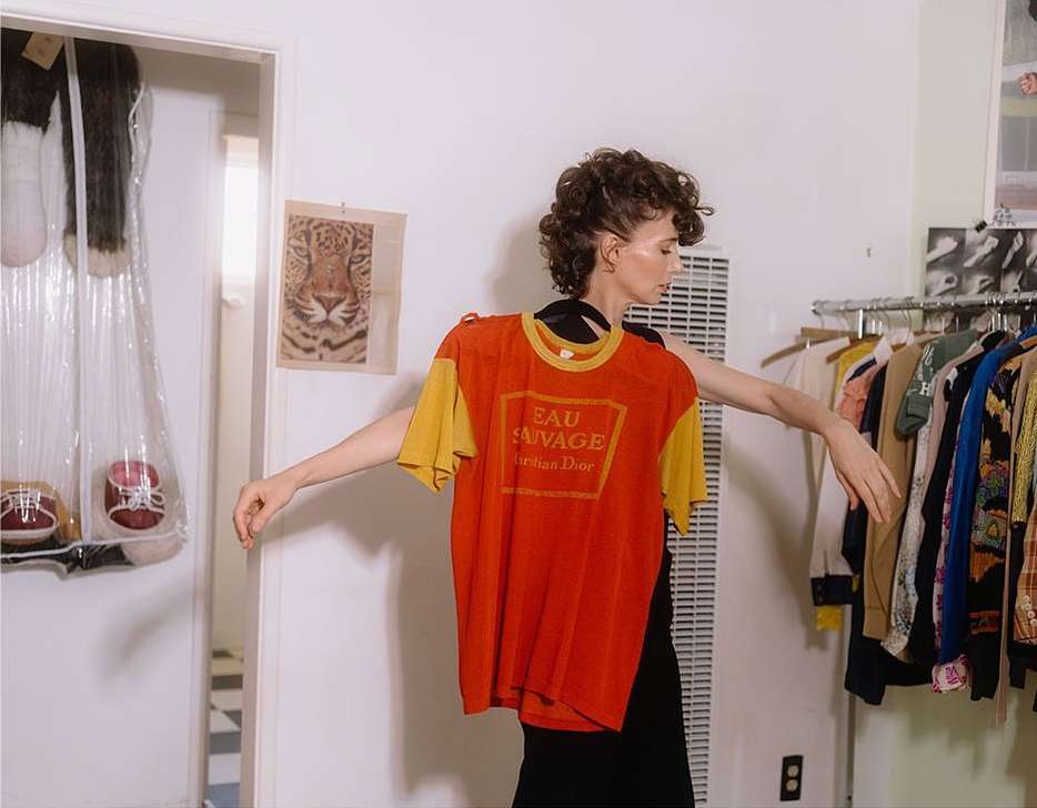 Adult female poses in an indoor setting, holding a vintage orange Eau Sauvage Dior t-shirt against a clothing rack.