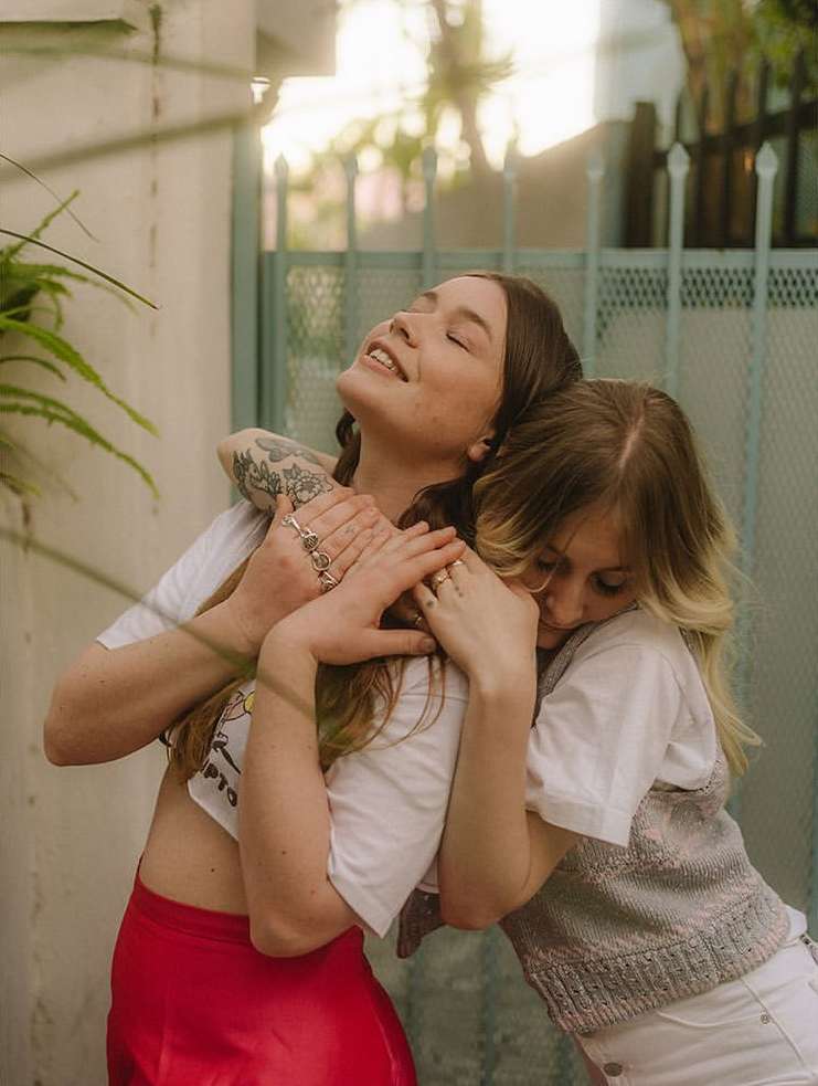 Two women embracing outdoors, one in a red skirt and white top, the other in a white top and gray vest, with plants and a wall behind them.