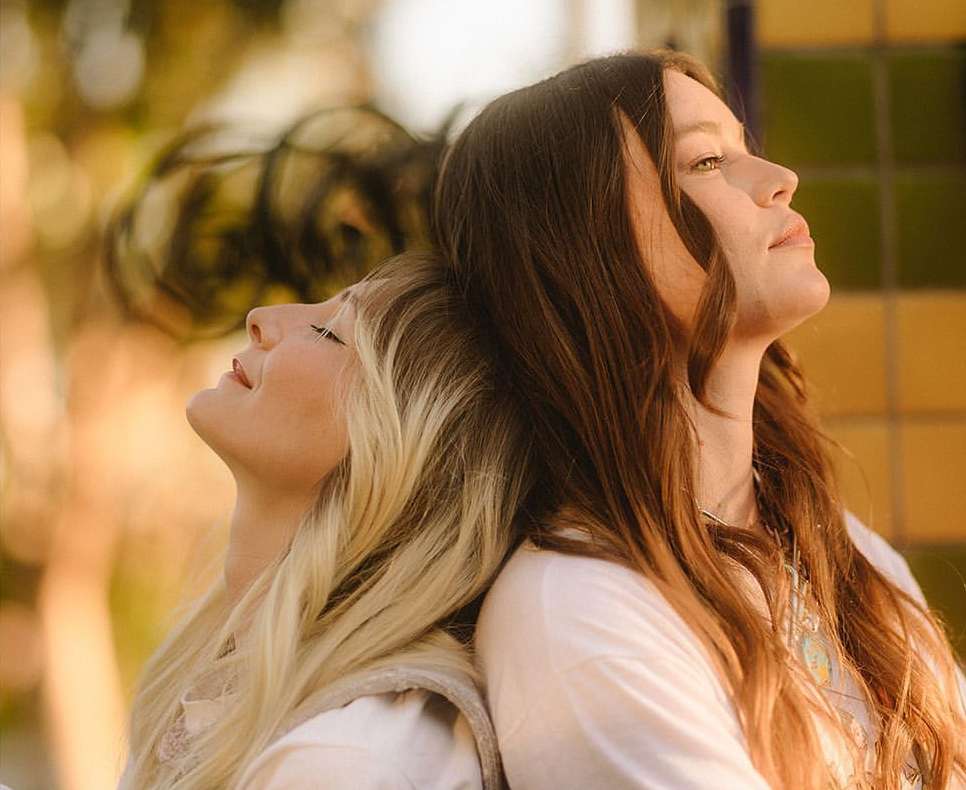 Two women with long hair lean back-to-back, basking in natural light with shallow depth of field. Portrait orientation in an outdoor setting.
