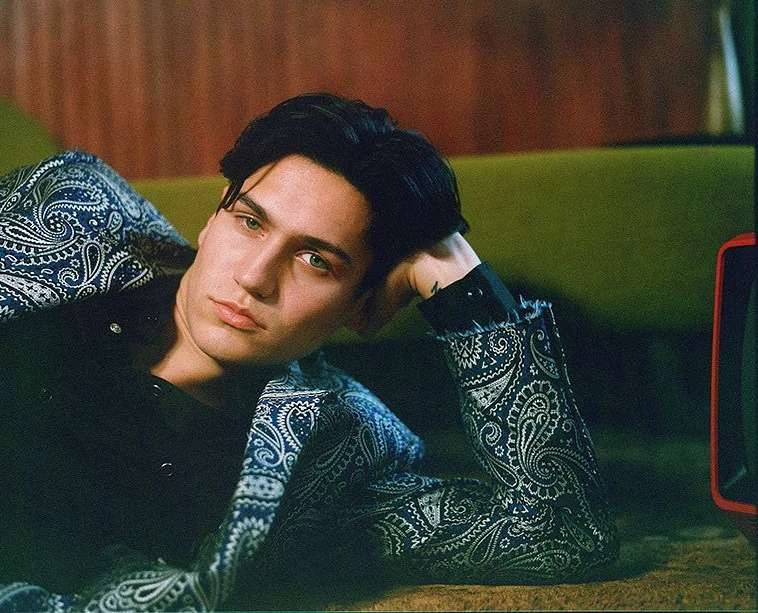 Portrait of a brunette man in a paisley suit reclining against a green sofa, looking off camera with a contemplative expression.