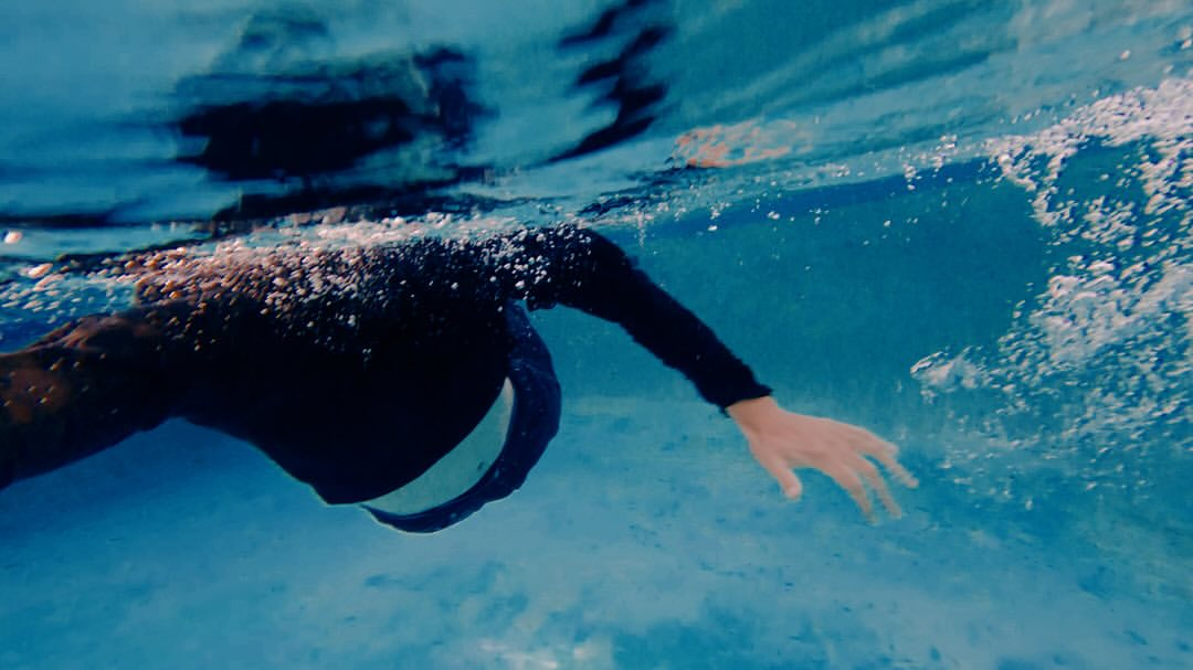 Underwater view of an adult swimming in a pool with blue tiles, wearing a black long-sleeve shirt and white stripe detail.