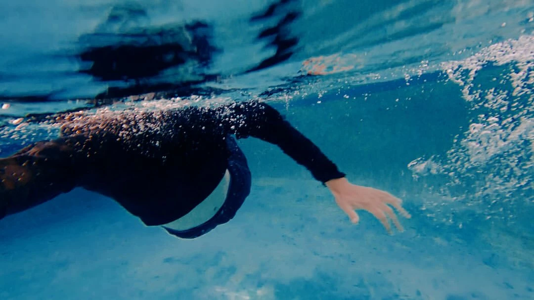 Underwater view of an adult swimming in a pool with blue tiles, wearing a black long-sleeve shirt and white stripe detail.