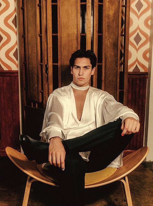 Young adult male poses in sheer white shirt and black pants, seated on a wooden chair in front of a paneled wood wall and patterned wallpaper.
