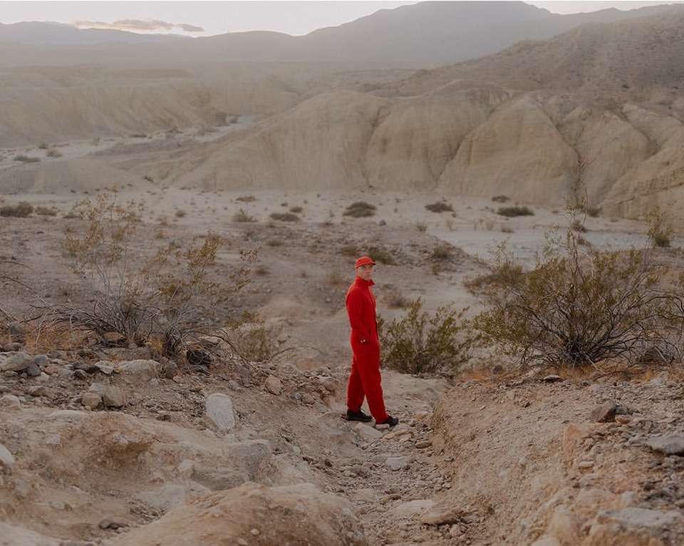 A person in a red jumpsuit and cap stands on a rocky, sandy path in a desert landscape with mountains and sparse vegetation, creating a solitary scene.