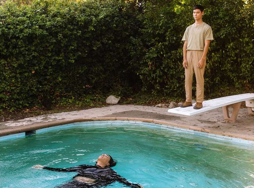 An adult male stands on a diving board next to a pool while an adult female floats in the water on a sunny day in an outdoor setting.