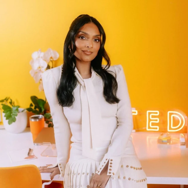 Portrait of a stylish woman in a cream-colored outfit posing in a bright studio with white desk and an illuminated neon sign.