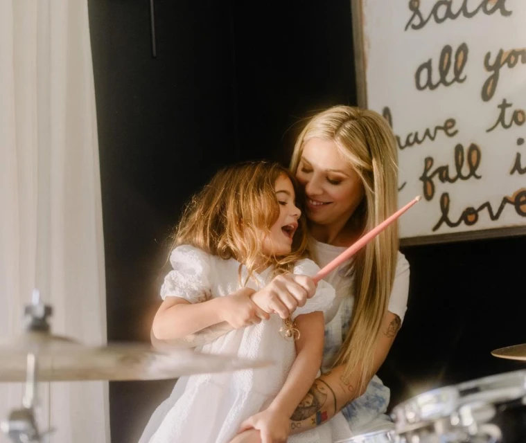 Blonde woman holds drum sticks while hugging her young daughter who wears a white dress next to a drum set indoors.