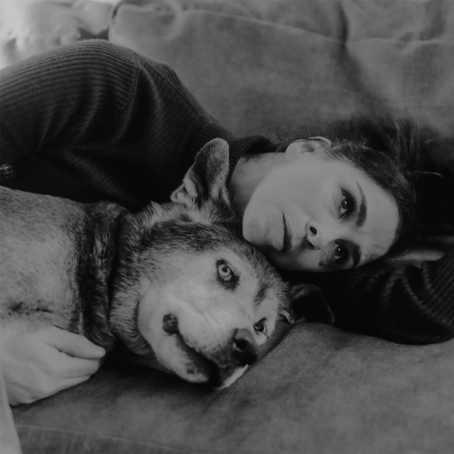 Black and white portrait of a woman with a wolf-like dog on a sofa, close together. The woman rests her head on the dog.