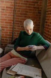 A person with short platinum blonde hair wearing a green turtleneck sweater seated on a couch looking at a smartphone.