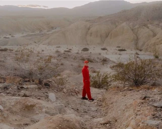 Person wearing a bright red jumpsuit and matching cap standing amidst arid desert hills and sparse scrubland.