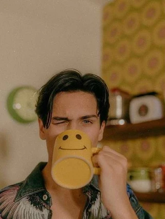 Young man drinking from a yellow smiley face mug in a kitchen with retro wallpaper, photographed by Elizabeth Weinberg.