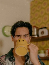 Young man drinking from a yellow smiley face mug in a kitchen with retro wallpaper, photographed by Elizabeth Weinberg.