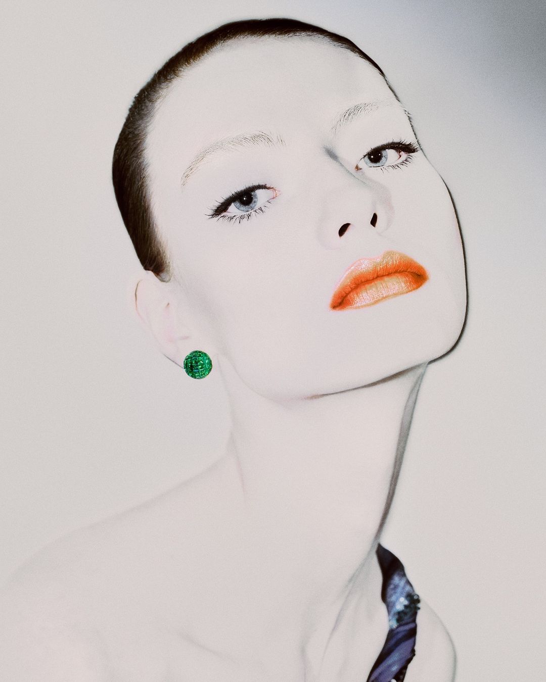 Close-up portrait of a fair-skinned adult female with short brunette hair, blue eyes, and coral lipstick, wearing a green round earring.