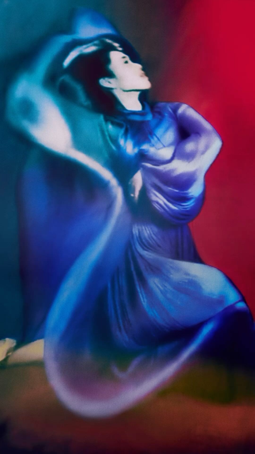 Woman in a flowing blue dress and matching hat poses indoors against a vibrant blue and red backdrop in a studio shot.