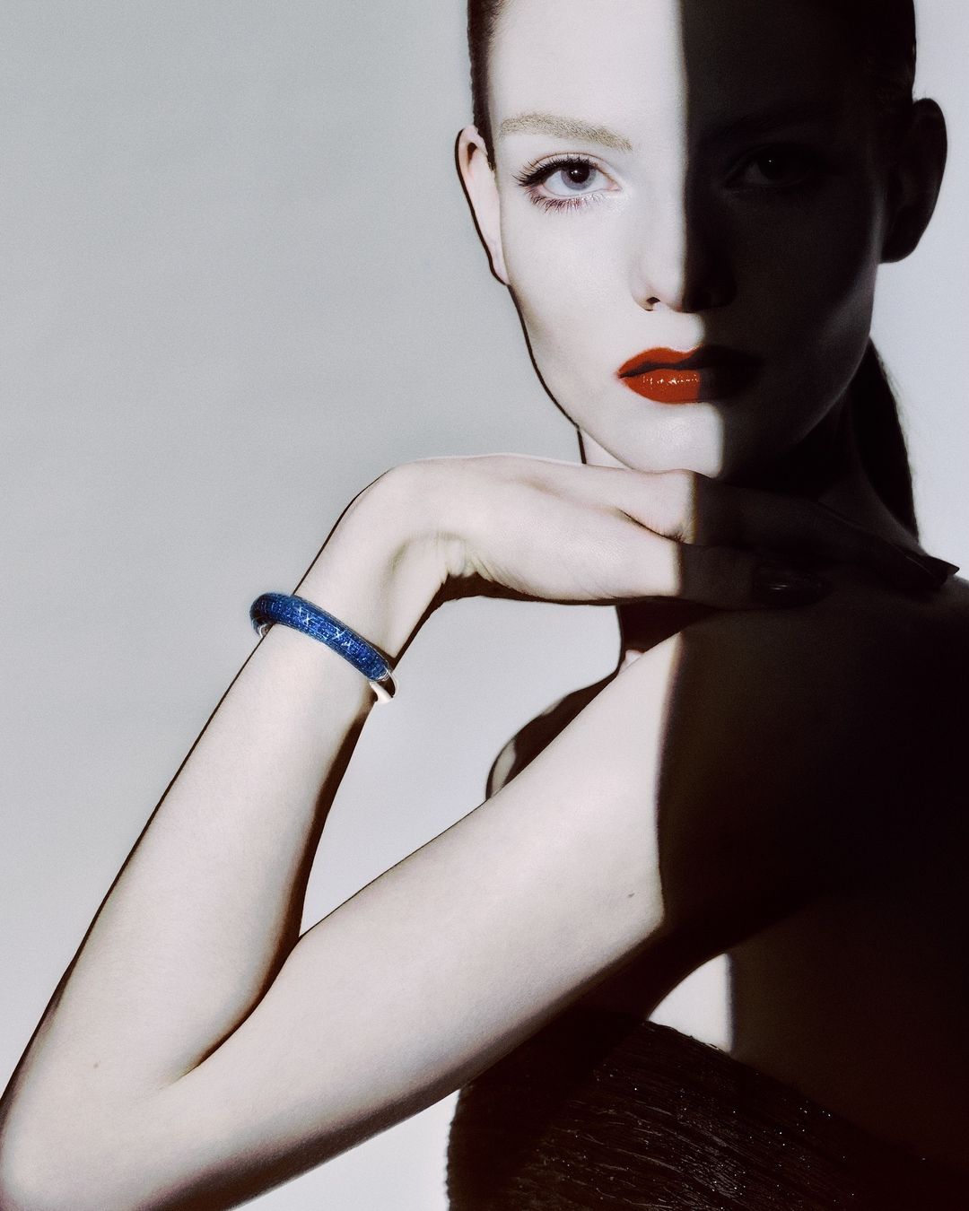 Close-up fashion shot of a fair-skinned woman wearing red lipstick and a sparkly blue bracelet in a studio setting with strong shadows.