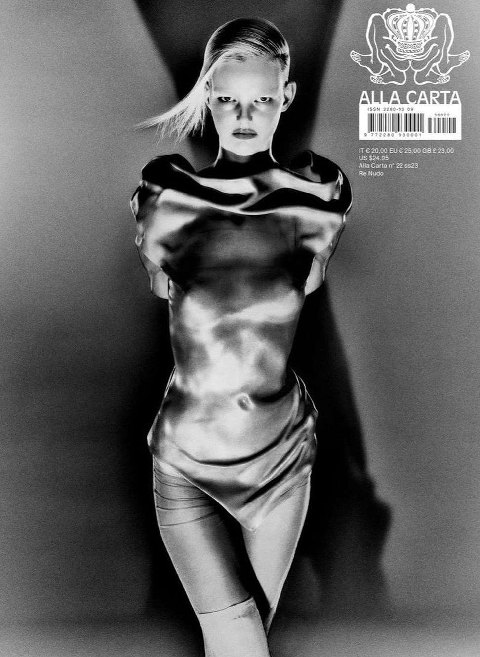 Monochrome studio portrait of a slim young woman in a silvery-toned outfit and light hair against a shadowy background with magazine graphics.