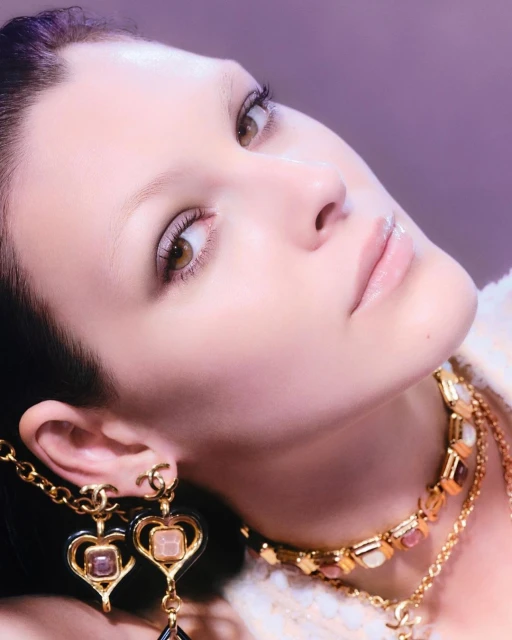 Close-up portrait of an adult woman wearing gold Chanel jewelry including earrings and a necklace, set against a soft purple background.