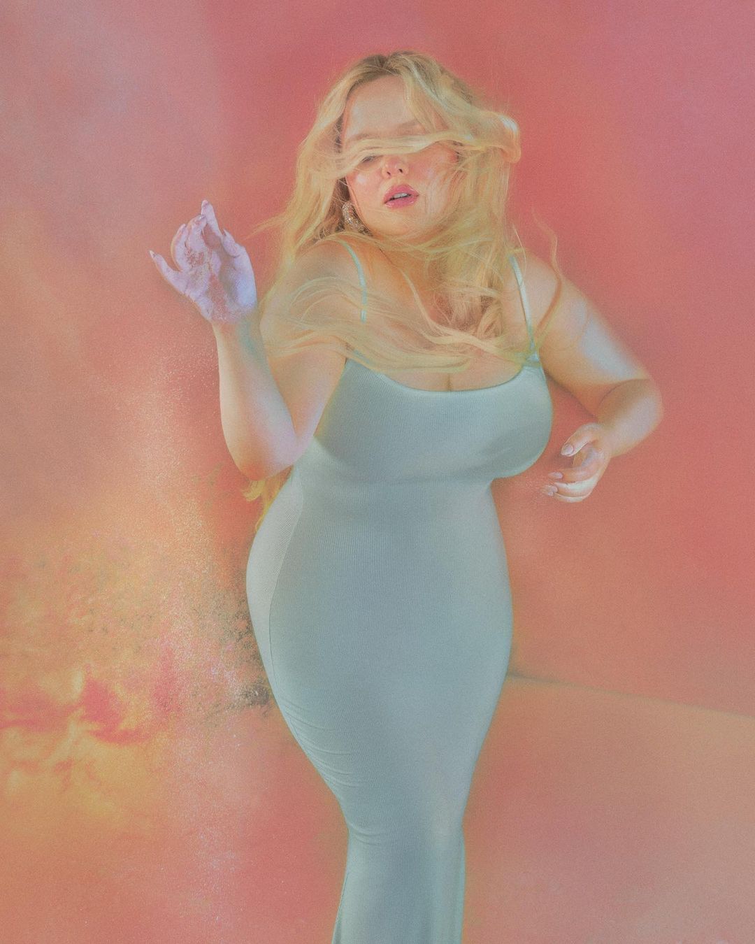 Blonde woman wearing a fitted pale green slip dress against a soft warm pink and orange gradient background