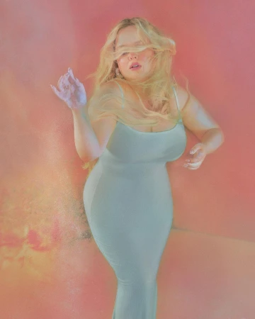 Blonde woman wearing a fitted pale green slip dress against a soft warm pink and orange gradient background