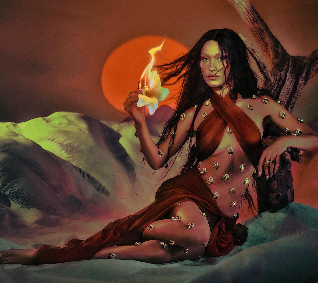 Woman with long dark hair in a sheer brown dress holding a burning flower in a desert landscape, with a sunset in the background.