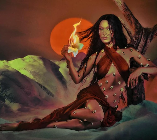 Woman with long dark hair in a sheer brown dress holding a burning flower in a desert landscape, with a sunset in the background.