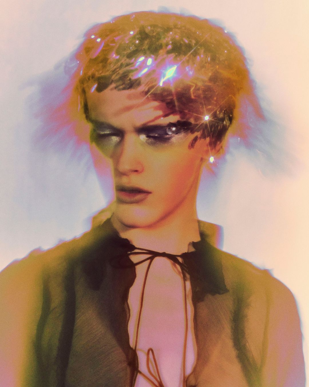 Close-up portrait of a model with sparkling hair, dramatic eye makeup, and an open black shirt against a soft, diffused background.