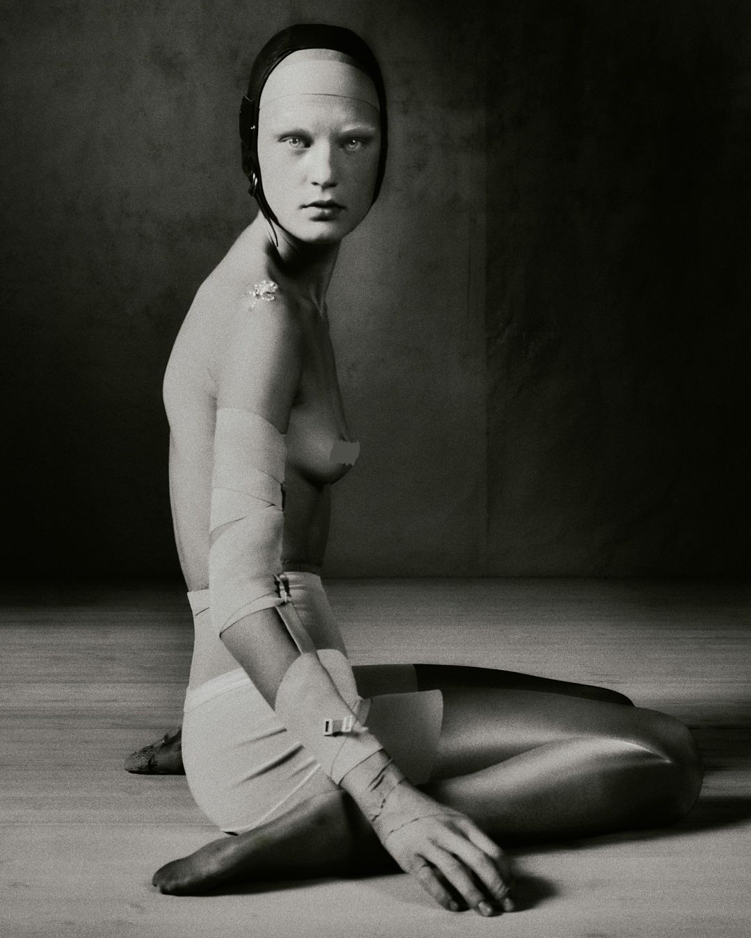 Portrait of a pale, bald adult female sitting on the floor with bandages and head covering, set against a dark and textured background.