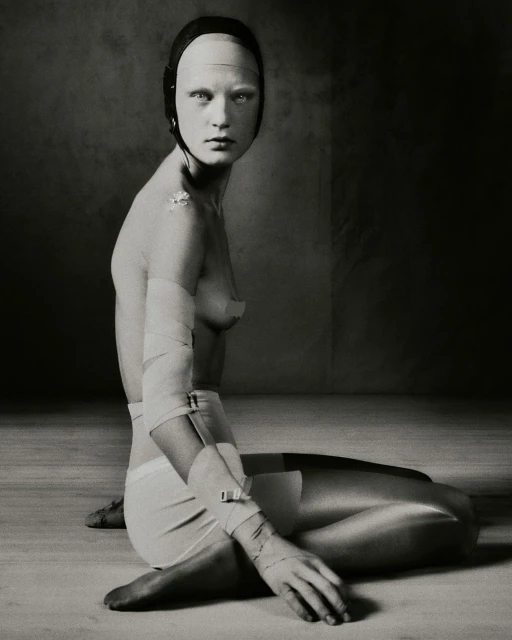 Portrait of a pale, bald adult female sitting on the floor with bandages and head covering, set against a dark and textured background.