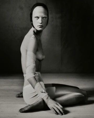 Portrait of a pale, bald adult female sitting on the floor with bandages and head covering, set against a dark and textured background.