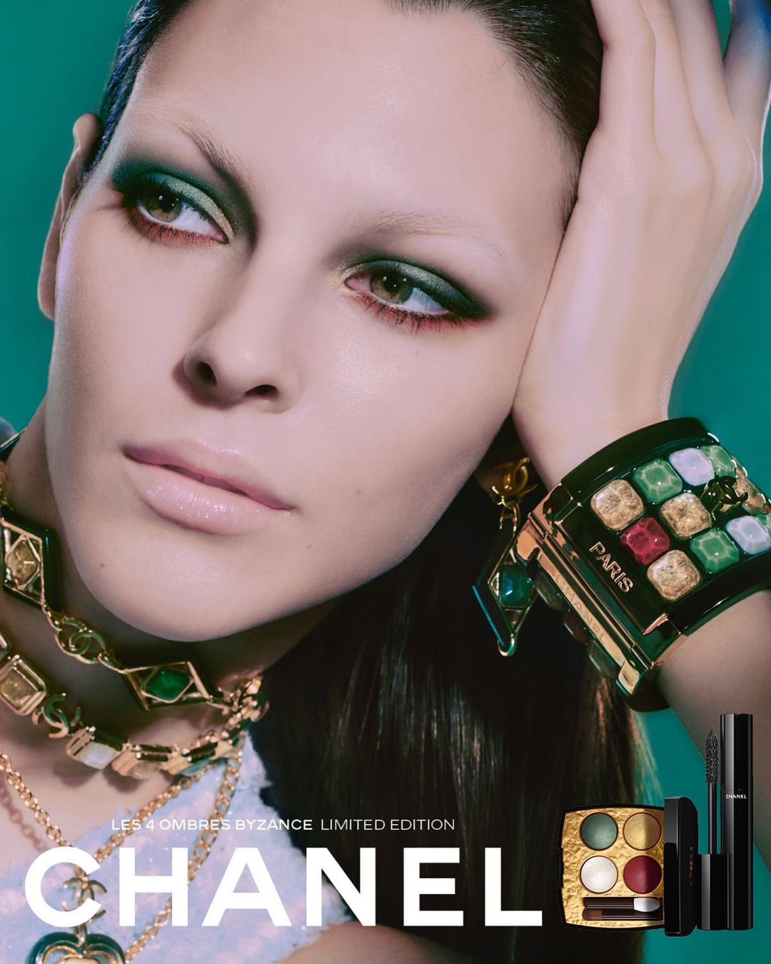 Close-up of a woman with green eye shadow, wearing layered gold necklaces and a large jeweled bracelet, promoting Chanel cosmetics.