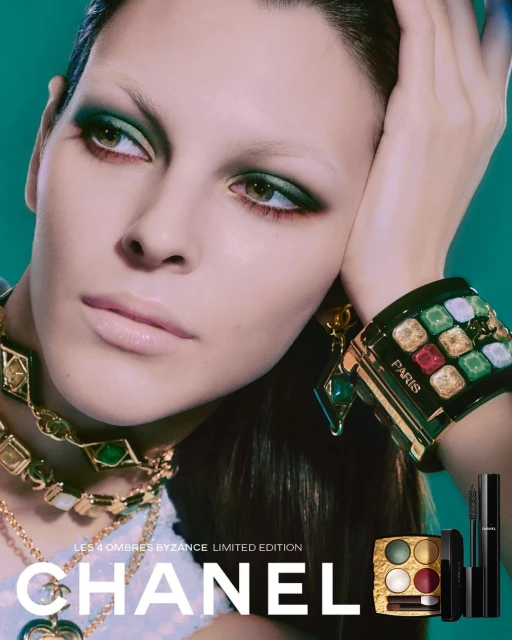 Close-up of a woman with green eye shadow, wearing layered gold necklaces and a large jeweled bracelet, promoting Chanel cosmetics.