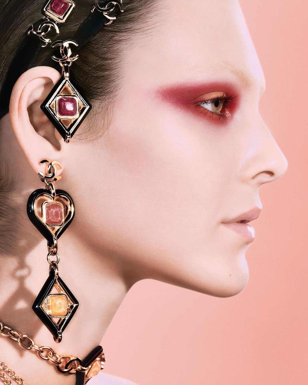 Profile shot of a woman with ruby eyeshadow and black and gold jewelry, soft peach background, fashion and beauty visual content.