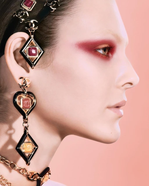 Profile shot of a woman with ruby eyeshadow and black and gold jewelry, soft peach background, fashion and beauty visual content.
