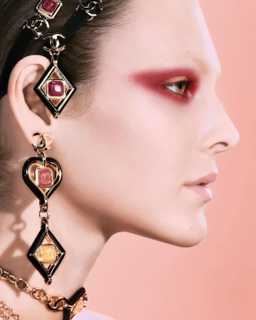 Profile shot of a woman with ruby eyeshadow and black and gold jewelry, soft peach background, fashion and beauty visual content.