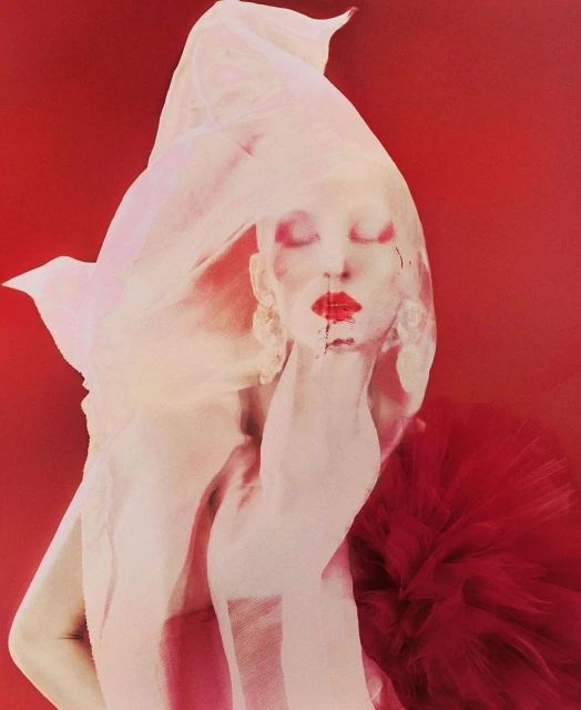 Artistic fashion portrait of a person with face paint and sheer fabric on a red background, showcasing creative beauty styling.