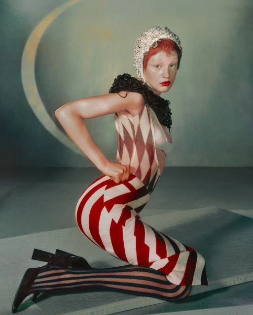 A woman in a harlequin-style outfit kneels indoors, featuring red hair, bright red lipstick, a textured collar and head covering.
