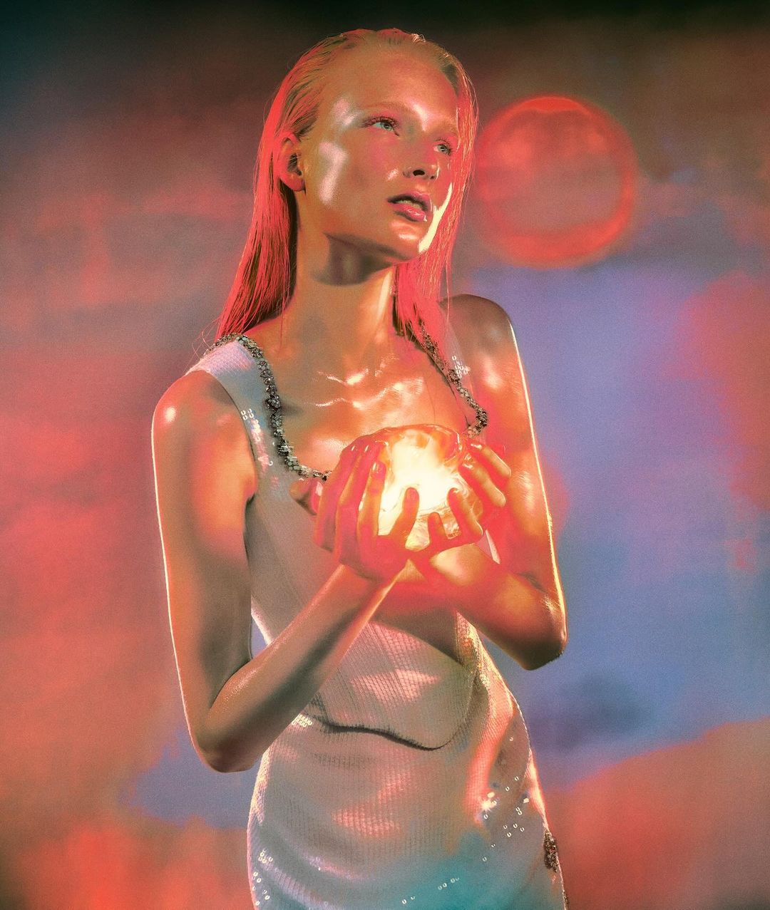 Portrait of a female model with long red hair, pale skin, wearing a white dress, holding a glowing orb in her hands against a hazy red background.