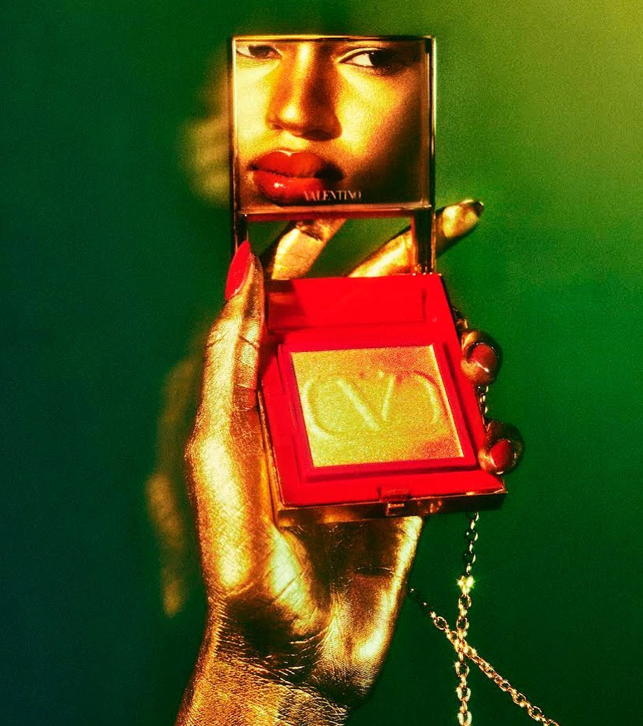Cosmetic product advertisement featuring a golden hand holding a Valentino blush compact, reflecting a face in the compact's mirror.