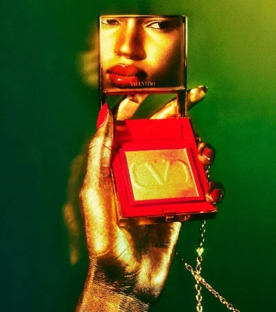 Cosmetic product advertisement featuring a golden hand holding a Valentino blush compact, reflecting a face in the compact's mirror.