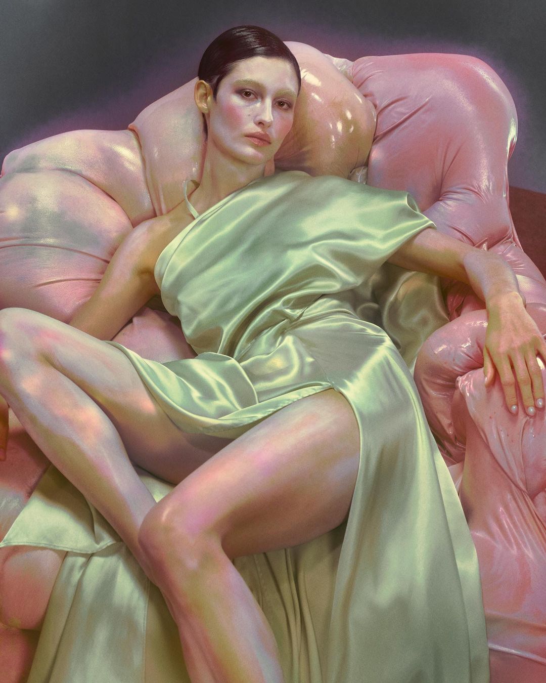 A woman in a pale green satin dress is seated on a pink, inflated-looking chair. The portrait-style shot features a soft lighting mood.