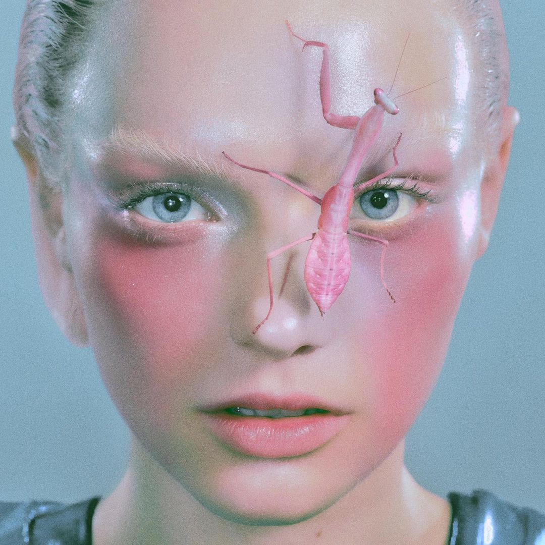Close-up portrait of a woman with light skin, blue eyes, and pink makeup, featuring a pink praying mantis on her forehead.