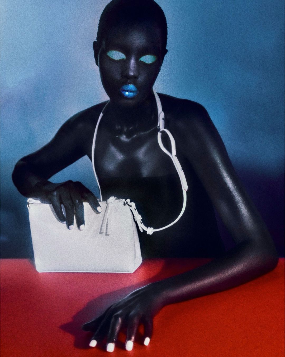 A dark-skinned adult woman is posing with a white purse on a red surface against a blue background in a studio-style portrait.
