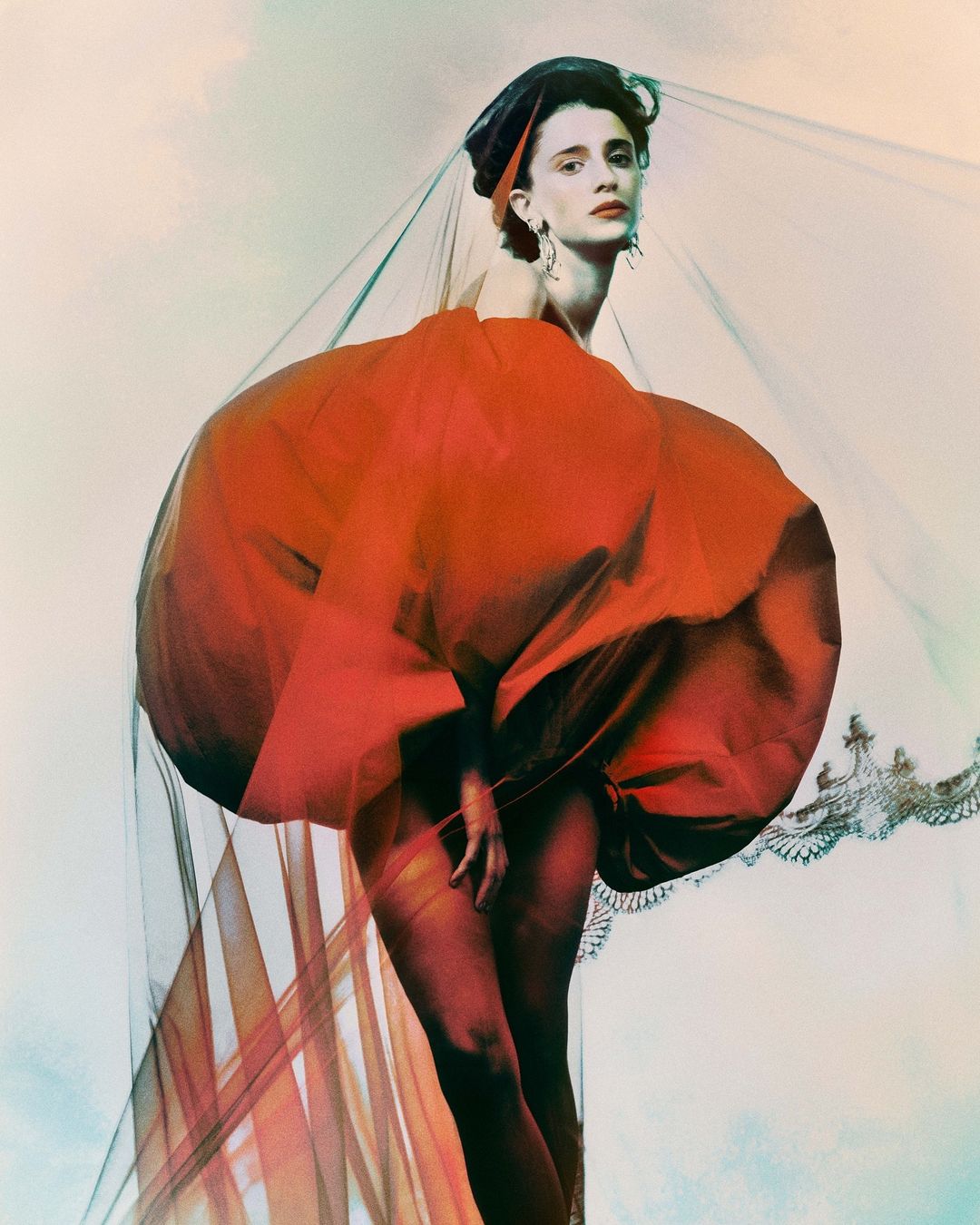 A fashion portrait features a woman in a large red gown with sheer draping and a headpiece, set against a soft, cloudy backdrop.