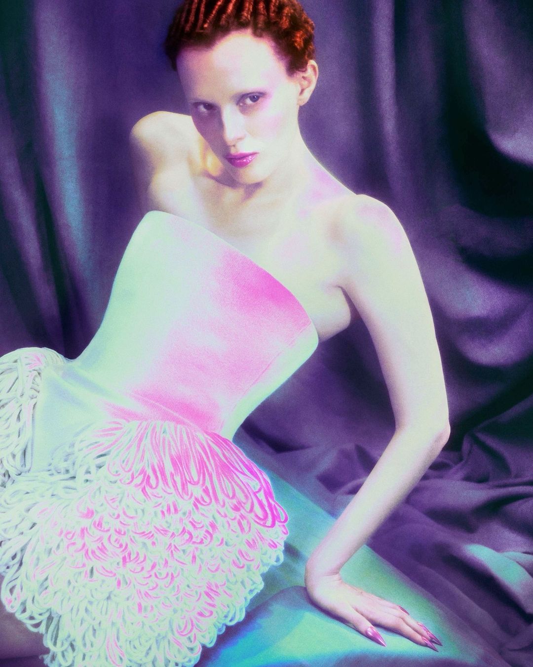 Adult female with red hair poses in a light pink strapless corset dress with fringe detail, set against a purple draped background.