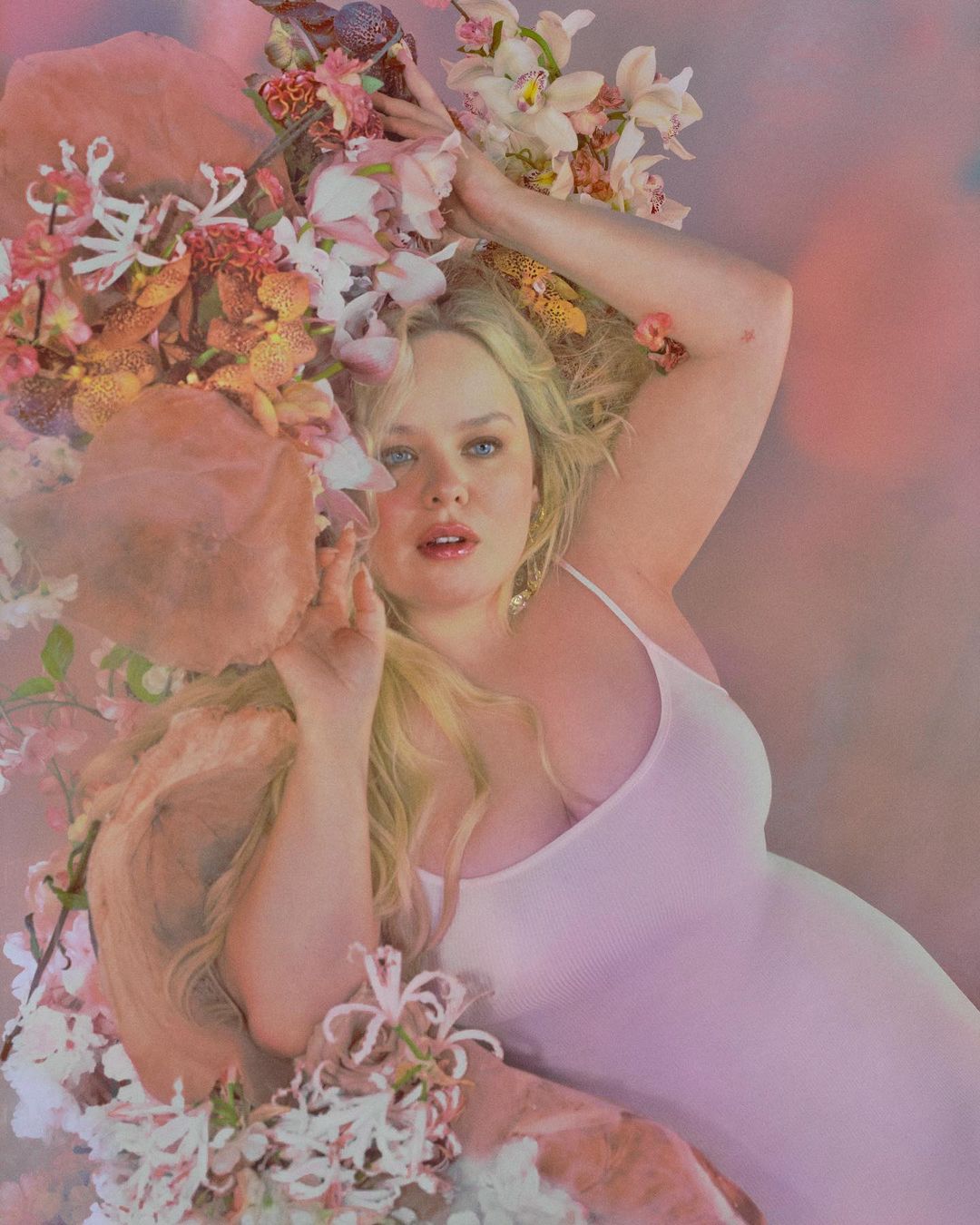 Portrait of an adult woman with blonde hair surrounded by flowers and wearing a white dress against a blurred, soft pink background.