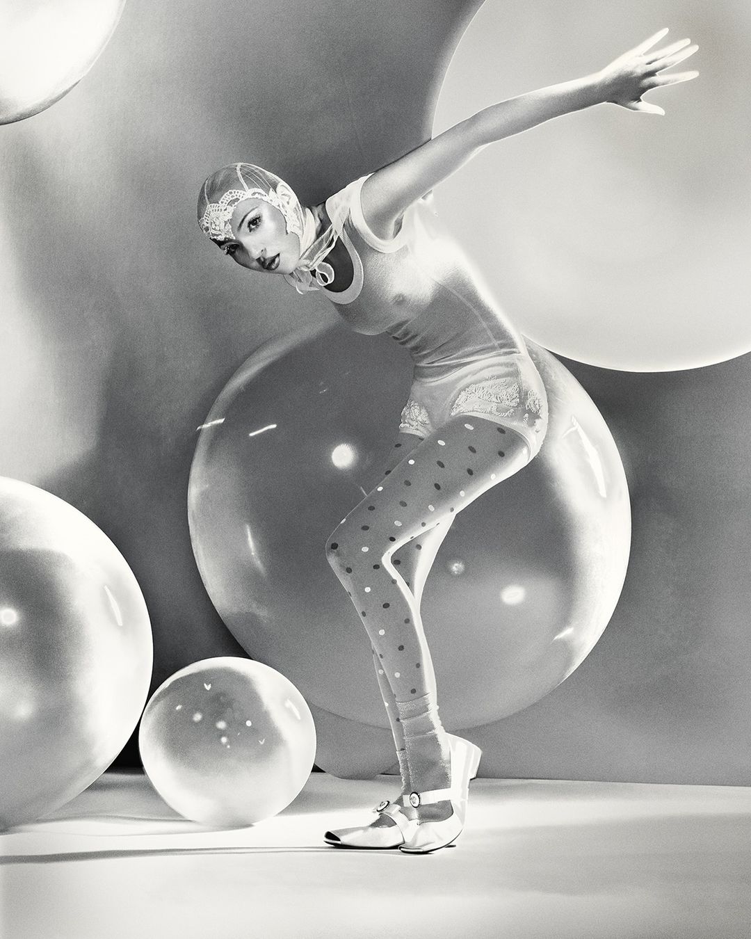 Woman in vintage-style bathing suit and polka-dot stockings poses among large, translucent spheres in a black and white studio portrait.