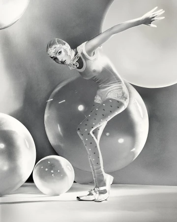 Woman in vintage-style bathing suit and polka-dot stockings poses among large, translucent spheres in a black and white studio portrait.