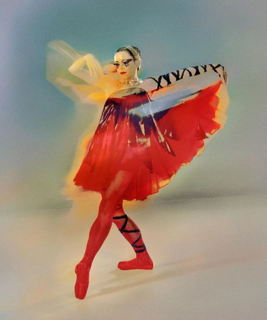 Dynamic photo of a ballet dancer in red attire performing a pose against a soft, diffused backdrop with blurred motion effect. 