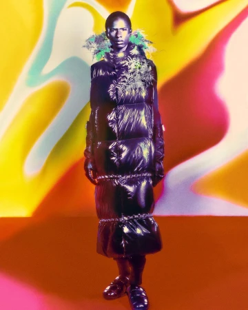 Black male model stands in a glossy black puffer coat with green floral accents, posing in a studio against a vibrant, abstract background.