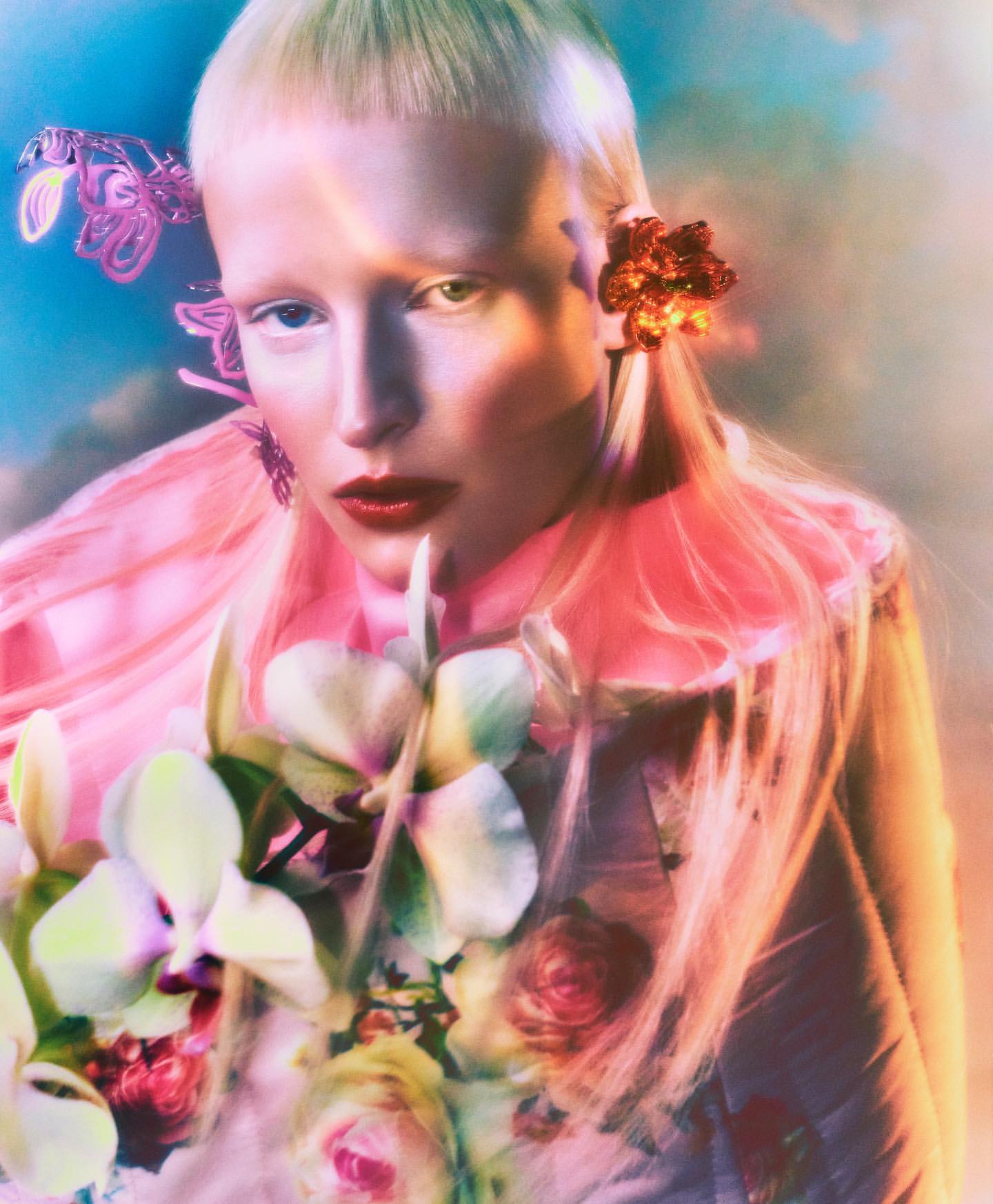 Dreamy portrait of a fair-skinned person with bright red lips, adorned with floral accents and set against a soft sky backdrop.